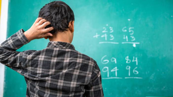 Confused kid scratching head to solve mathematics problem on board at classroom - concept of intelligence, trouble learning and education